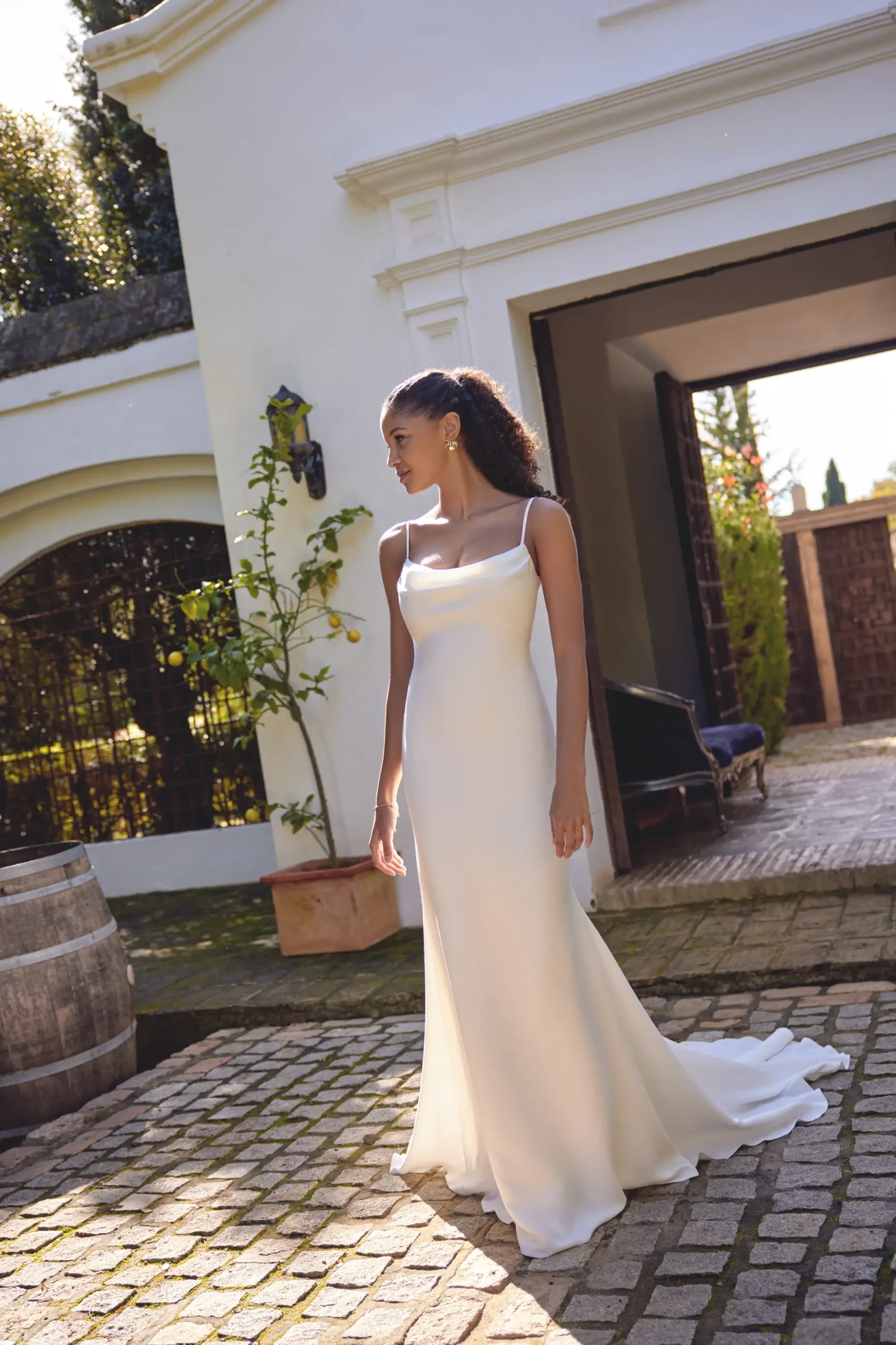 A woman in a white dress stands on a cobblestone path outside a building, with greenery and decor in the background.