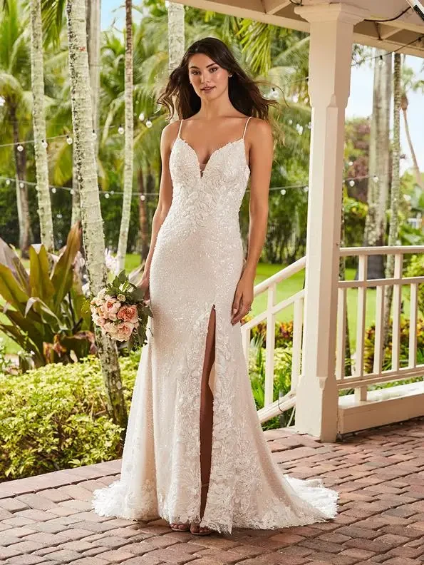 Model is wearing a sequined gown with lace appliqués, a daring front slit, chapel-length train, thin beaded straps, and a low keyhole back.
