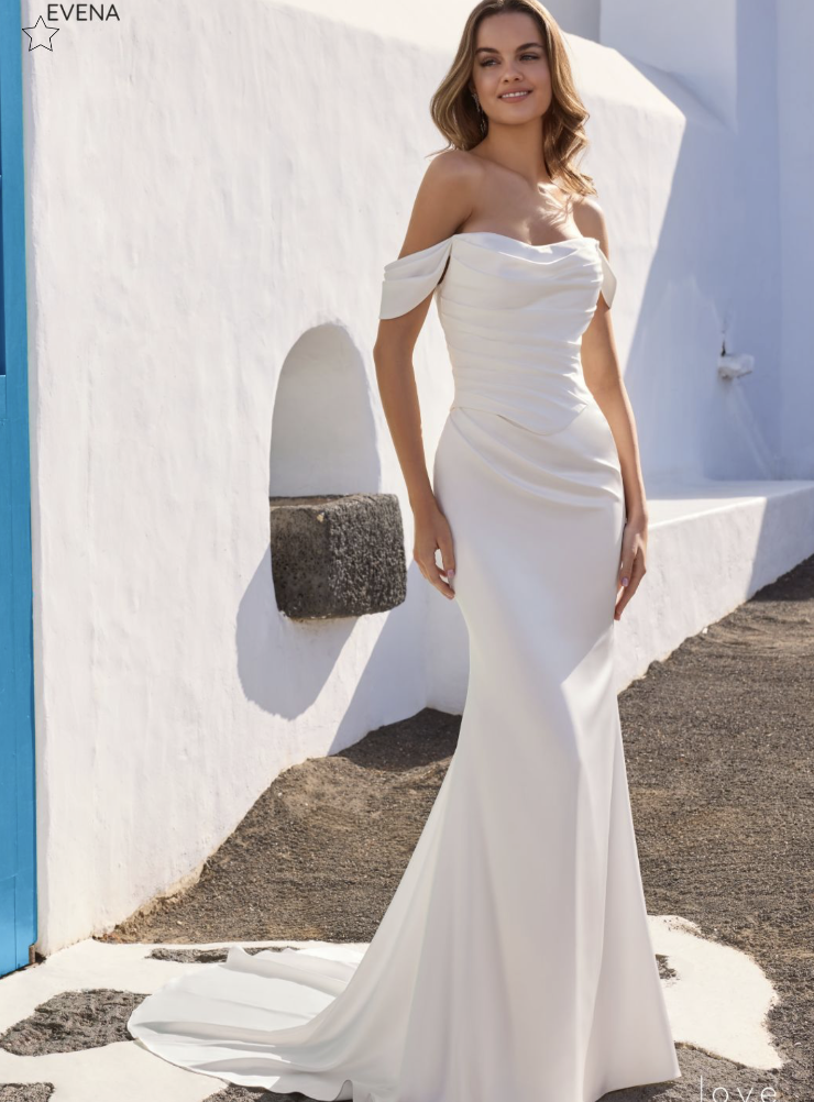 A woman in an elegant, off-the-shoulder white gown stands outdoors against a white building with blue accents. The scene is sunny and idyllic.