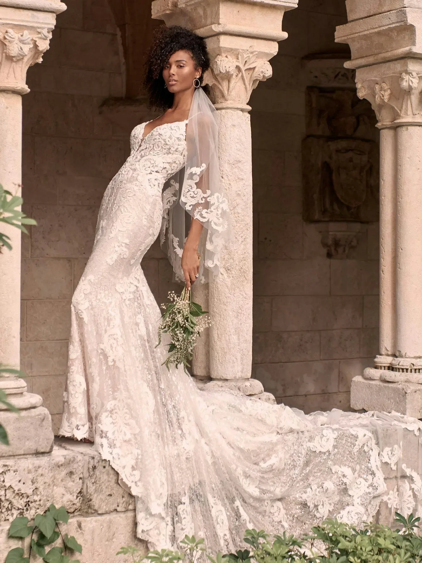 A bride in an elegant lace gown with a long train poses against stone columns. She holds a small bouquet, exuding a serene and graceful vibe.