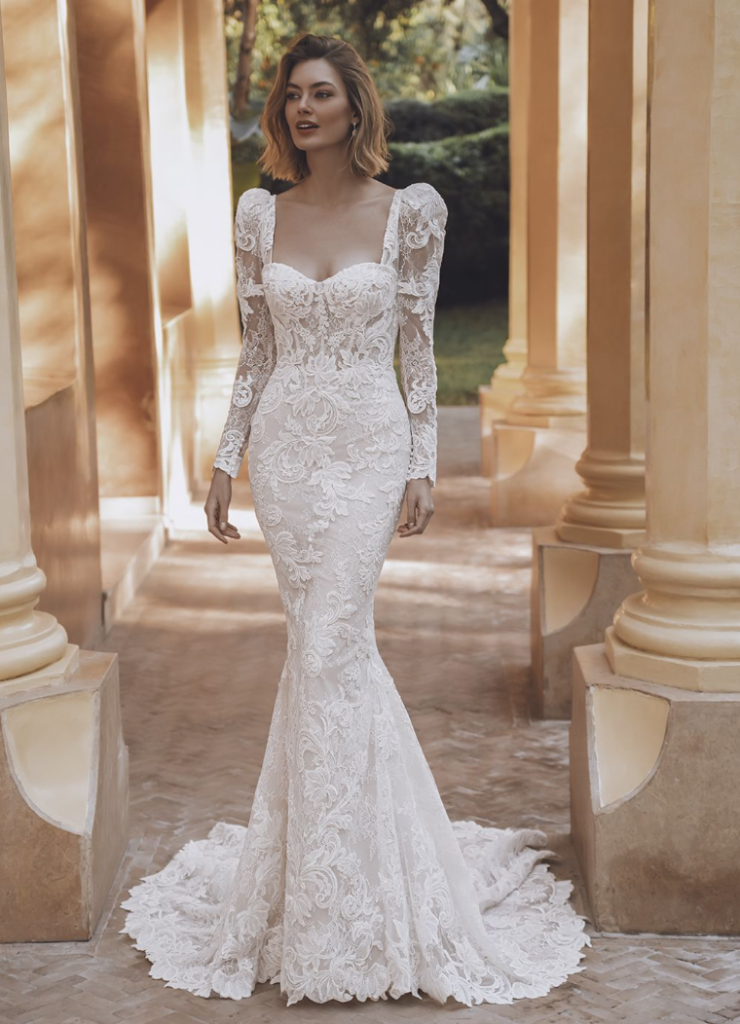 Bride in elegant ivory lace gown with long sleeves and a train stands between ornate columns. Sunshine casts soft shadows; a serene, romantic ambiance.