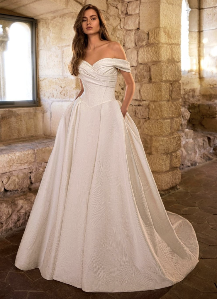 A woman wearing a beautiful off-the-shoulder white wedding gown, standing in a stone room with large windows.
