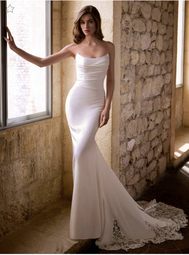 A woman in a strapless, fitted white dress standing near a stone wall with large windows.