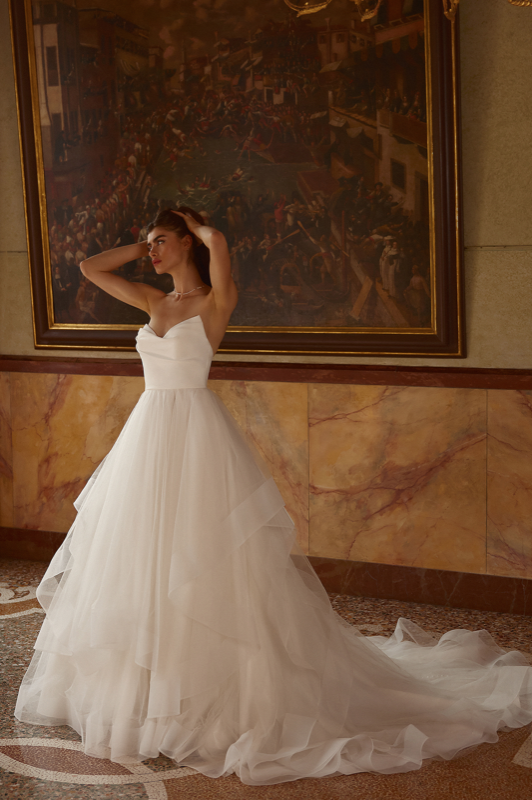 A woman in a white wedding dress poses elegantly in a room with ornate decor and dramatic artwork on the wall.