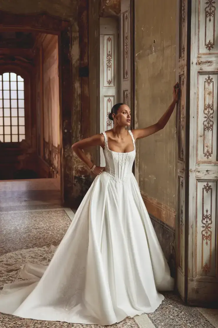 A woman in a fitted white gown with a flowing skirt stands elegantly against a wall, showcasing an ornate interior with large windows in the background.