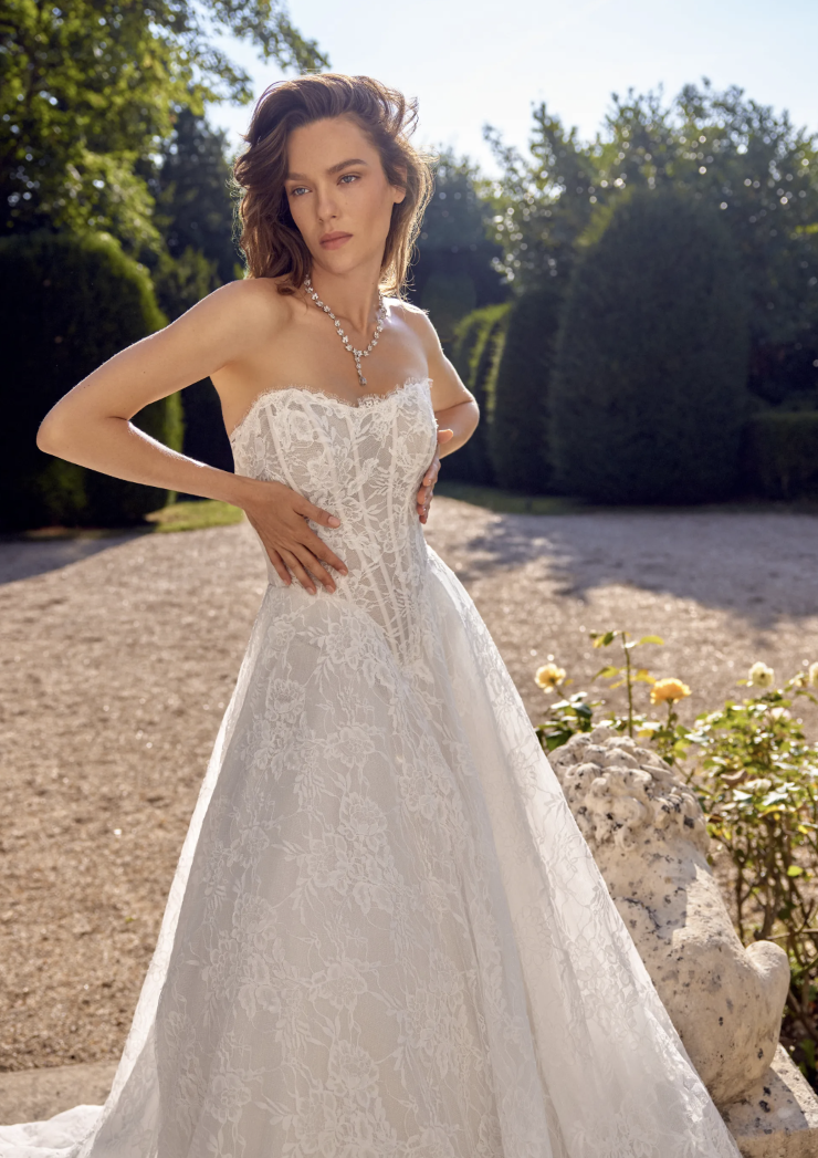 A woman wearing a strapless white lace wedding dress poses outdoors in a garden setting, with greenery and sunlight in the background.
