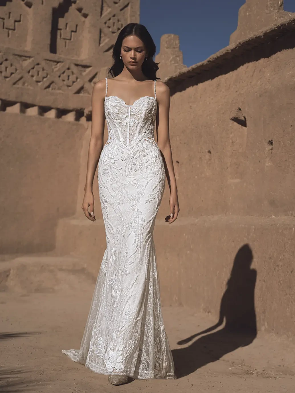 A model wearing an elegant white wedding dress with intricate detailing, walking in a desert-like setting against a backdrop of textured earth walls.